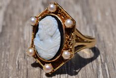 The Harper Antique Filigree Cameo Ring finished with six Pearls surrounding the center. The center is a hand carved cameo. Surrounding the center between the pearls is an intricate filigree pattern. The ring is crafted in 14 karat yellow gold and is very comfortable to wear. This ring is currently a finger size 6 3/4, but can be adjusted in size. Each piece has been hand selected and meticulously identified and graded by a Graduate Gemologist who has been awarded a degree by the Gemological Inst White Cameo Wedding Rings, Classic Wedding Filigree Ring With Cabochon, White Cameo Rings For Wedding, Heirloom Filigree Ring With Cabochon For Wedding, Heirloom Filigree Wedding Ring With Cabochon, Elegant Oval Filigree Ring With Intaglio, Elegant Carved White Rings, Antique Carved Filigree Ring For Wedding, Elegant White Carved Rings