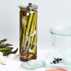 asparagus and other vegetables in a glass jar next to a measuring cup on a white surface