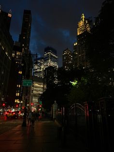 people are walking down the sidewalk in front of tall buildings at night time with lights on