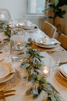 the table is set with white plates and silverware, candles and greenery on it