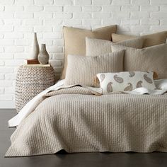 a bed with pillows and blankets on it in front of a white brick wall next to a basket