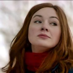 a woman with long red hair wearing a blue coat and a red scarf looks into the distance