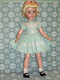 a doll with blonde hair wearing a blue dress and black shoes is standing in front of a wall