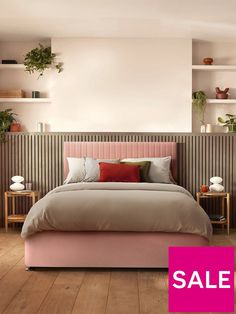 a bed with pink sheets and pillows in a bedroom next to some shelves filled with potted plants