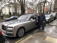 two men standing next to a silver rolls royce parked on the side of a road