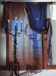 two candles are lit in front of a window with blue drapes and lace curtains