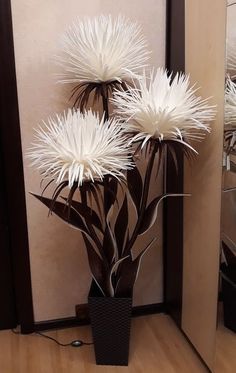 three white flowers in a black vase on the floor next to a mirror and wall