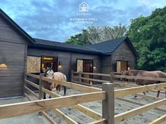 two horses are standing in the dirt near a wooden fence and some barn buildings with lights on them