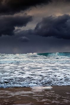 the sky is filled with dark clouds over the ocean and waves crashing on the beach