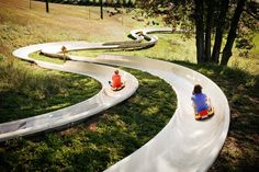 two children are riding down a hill on their toy cars in the park with trees and grass behind them