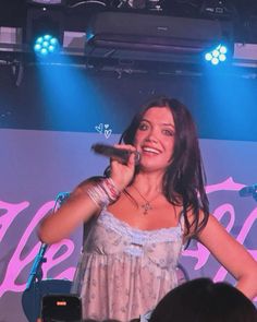 a woman holding a microphone in front of a stage with lights on it and people watching