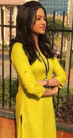 a woman in a yellow dress standing by a fence with her arms crossed and looking off to the side