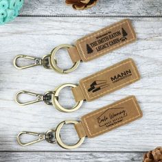 three personalized leather keychains are shown on a white wooden table with pine cones