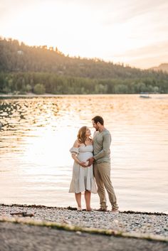 a man and woman standing next to each other on the shore of a lake at sunset