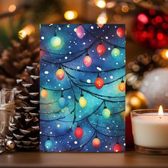 a christmas card with lights on it next to a lit candle and pine cones in the background