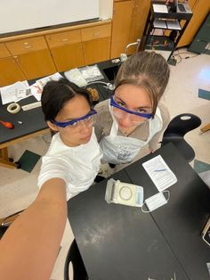 two girls wearing safety goggles while looking at something on the table in front of them