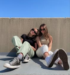 two young women sitting on the ground with their legs crossed and one is wearing sunglasses