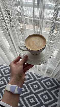 a person holding a cup of coffee in front of a window with sheer curtains on it