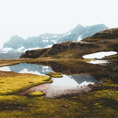 a small pond in the middle of a grassy field with mountains in the background and snow on the tops