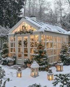 a small white house covered in snow with christmas lights on the windows and trees around it