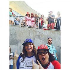 two people wearing baseball caps standing next to each other in front of a crowd with their thumbs up