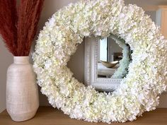 a white flower wreath sitting on top of a wooden table next to a vase filled with flowers