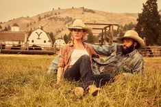 a man and woman sitting on the grass in front of a farm with mountains behind them