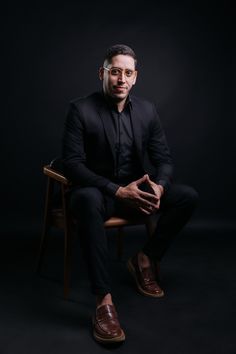 a man sitting on top of a wooden chair in front of a black background wearing glasses