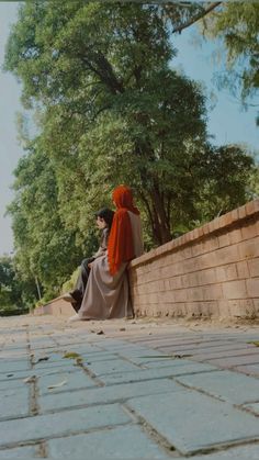 two people sitting on the side of a brick wall looking at each other and trees in the background