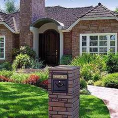 a brick mailbox in front of a house with grass and flowers around the yard