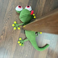 a crocheted green lizard laying on the floor next to a wooden table with yellow flowers