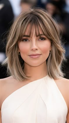 A layered bob with curtain bangs is perfect for adding movement and volume to medium-length hair. The warm blonde balayage highlights are perfect for adding dimension. This style suits round and heart-shaped faces, framing the face beautifully. The background shows a high-profile event, highlighting the cut’s sophisticated appeal. Chin Length Layers Medium Hair, Layered Hair Short With Curtain Bangs, Short Hair Bangs Heart Face, Layered Bob With Bangs Straight Hair, Flicky Bob Hairstyles, Medium Bob With Highlights, Short Hair Cut With Face Frame Bangs, Short Fall Hair With Bangs, Blonde Highlights Framing Face