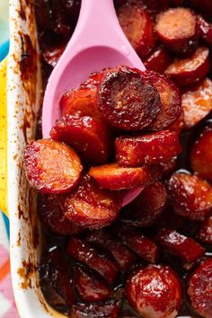 a pink spoon is in a dish full of cooked sausages