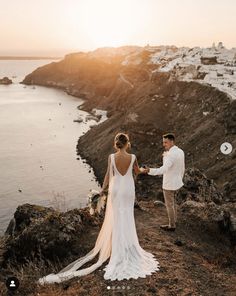 a bride and groom holding hands on top of a cliff overlooking the ocean at sunset