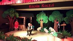 people are sitting on stage in front of trees and fake animals at the children's theater