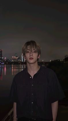 a young man standing in front of a body of water with the city lights behind him