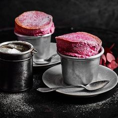 two desserts with pink frosting in metal cups and spoons on a black table