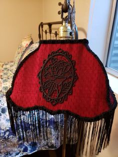 a red lamp shade with black fringes and a rose embroidered on the top, sitting on a bed