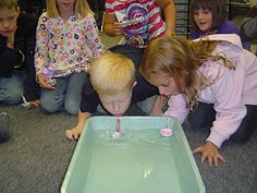 children are gathered around a water fountain in the middle of an office building while one child blows out a candle on it