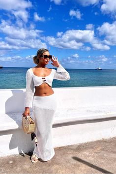 a woman standing next to the ocean with her hand on her hip and wearing sunglasses