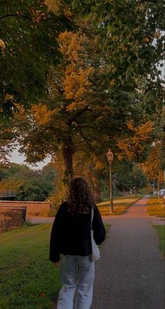 a woman walking down a sidewalk next to a tree filled park with lots of leaves