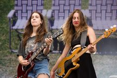 two young women playing guitars on stage at an outdoor music festival, one with long hair and the other without