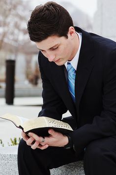 a man in a suit sitting on a bench reading a book and looking at it