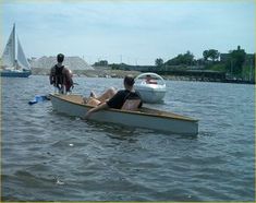 two people on a small boat in the water with sailboats and one person standing next to it