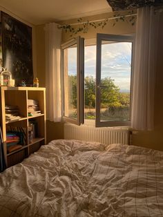 an unmade bed sitting in front of a window next to a bookshelf