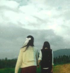 two people walking down a dirt road in front of a field with grass and trees