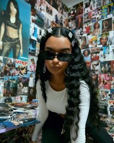 a woman with long black hair wearing sunglasses and sitting on the floor in front of a wall full of posters