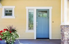 a blue front door on a yellow house with flowers in the foreground and white trim