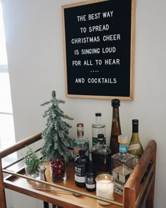 a wooden table topped with bottles of alcohol next to a chalkboard that says the best way to spread christmas cheer is singing loud for all to hear and cocktails