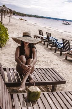 a woman in a straw hat sitting on a bench at the beach with a coconut